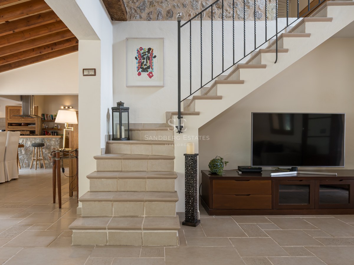 Vue intérieure montrant un escalier en pierre, un meuble TV en bois et un coin repas avec des poutres apparentes.