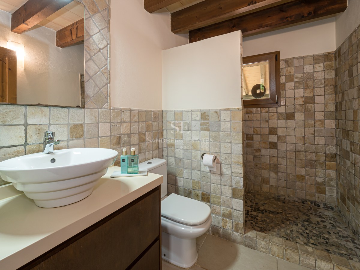 Salle de bain rustique avec murs en pierre, poutres apparentes, vasque blanche et douche au sol en galets.