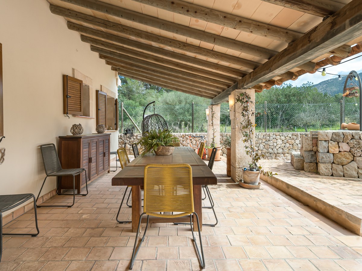 Terrasse couverte avec table en bois, chaises, murs en pierre et plafond à poutres avec vue sur la campagne.