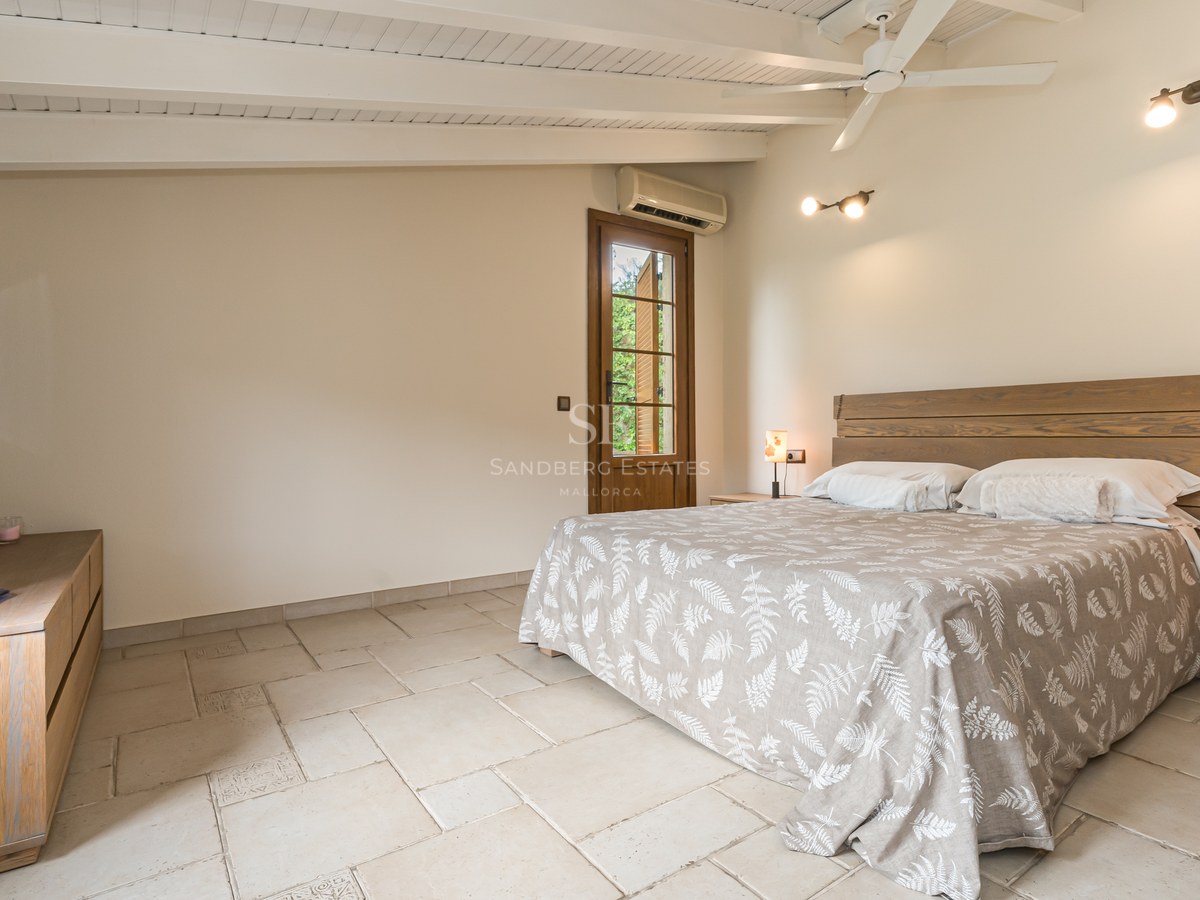 Chambre avec lit en bois, plafond à poutres blanches, sol en pierre et grandes fenêtres.