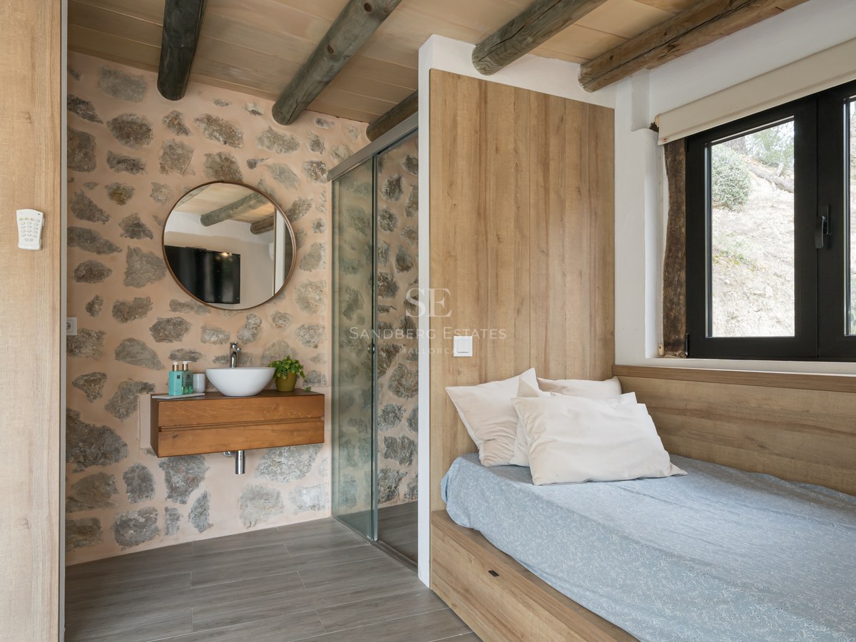 Chambre avec mur en pierre apparente, poutres en bois, lit intégré et coin vasque ouvert.