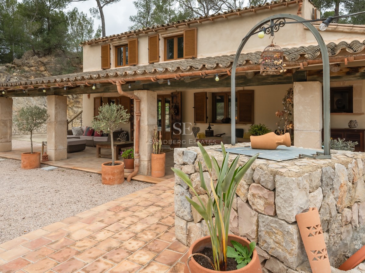 Façade d'une maison rustique avec porche couvert, volets en bois et un puits en pierre au premier plan.