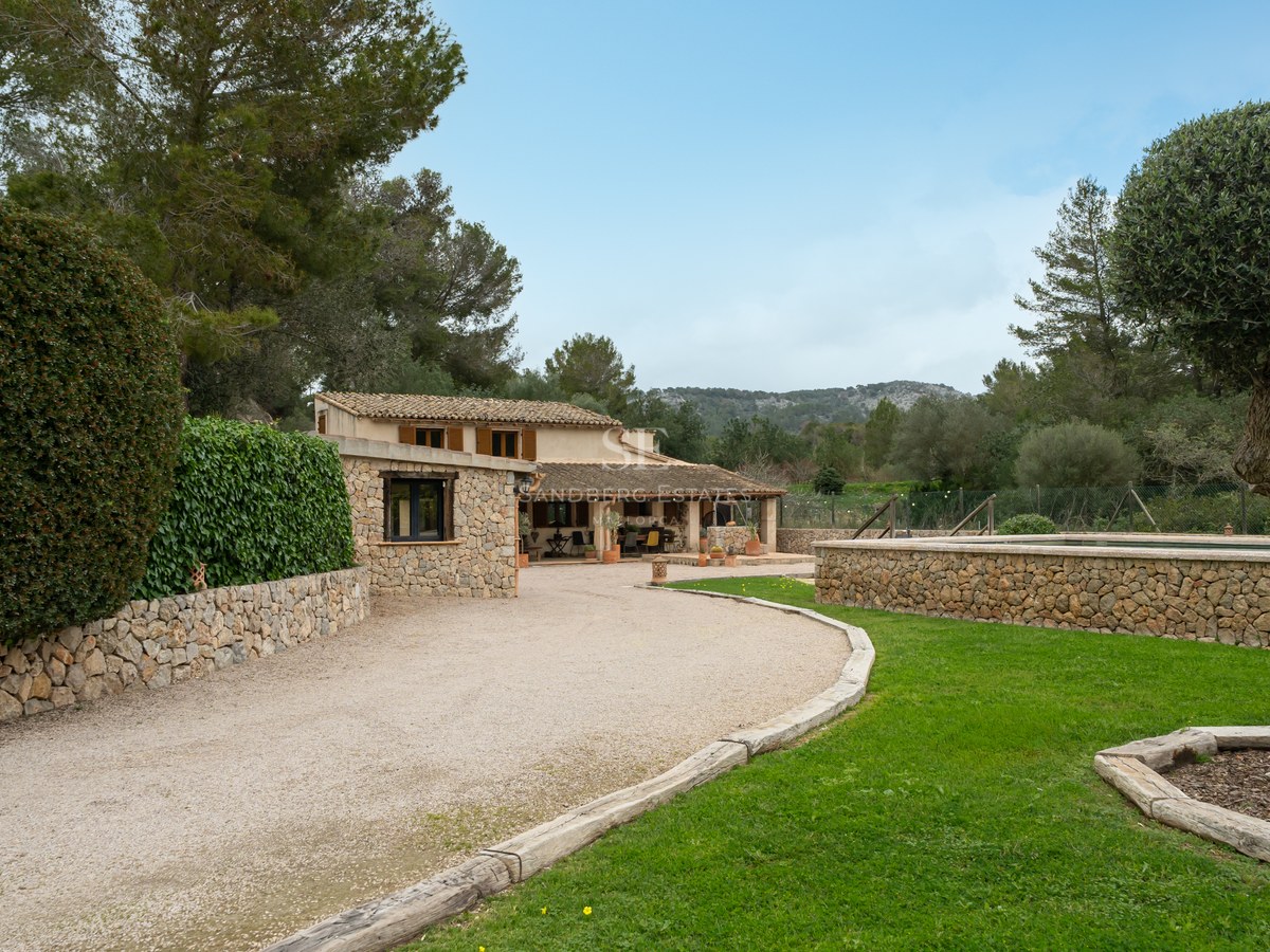 Maison en pierre rustique avec allée en gravier, pelouse verte et piscine en pierre surélevée sous un ciel clair.
