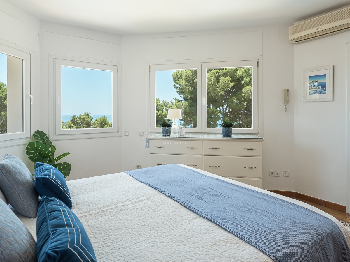 Chambre blanche lumineuse avec de grandes fenêtres vue mer, literie bleue et porte vers un balcon ensoleillé.