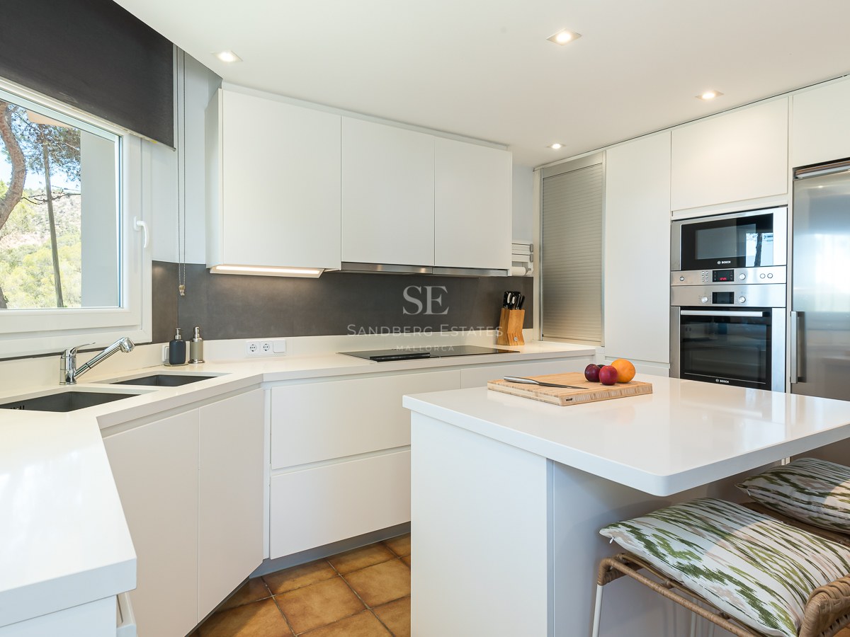 Cuisine moderne lumineuse avec placards blancs, appareils en inox, îlot central et vue sur la forêt par la fenêtre.
