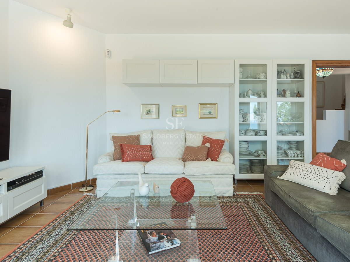 Salon lumineux avec canapés, table basse en verre sur tapis orné, grand téléviseur et sol en terre cuite.
