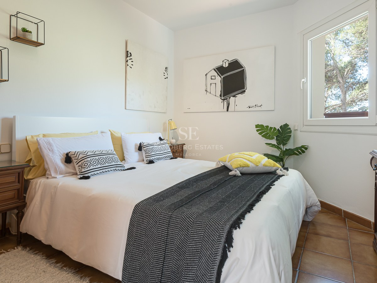 Chambre moderne avec lit blanc, coussins à motifs, tables de chevet en bois et sol en carrelage sous lumière naturelle.