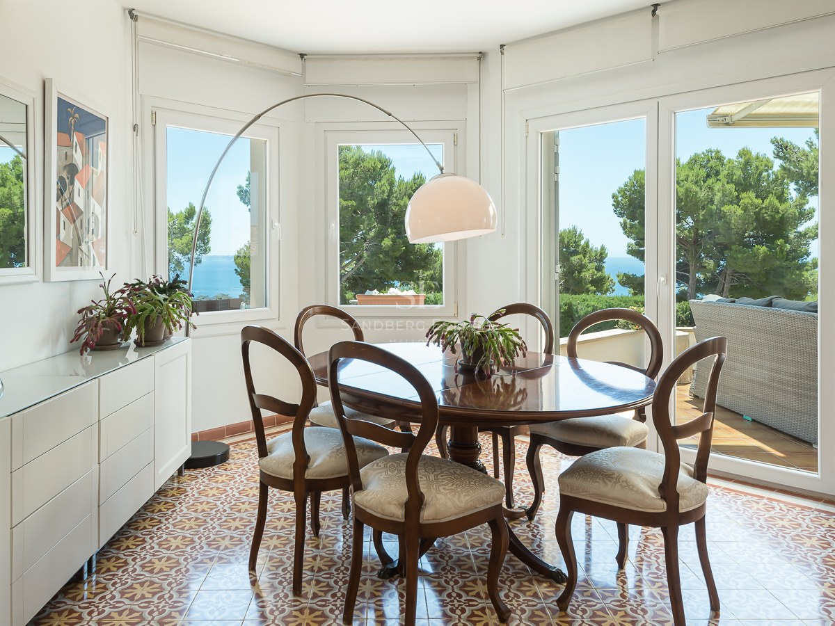 Salle à manger lumineuse avec table ronde en bois, sol en carreaux de ciment et grandes fenêtres donnant sur la mer.