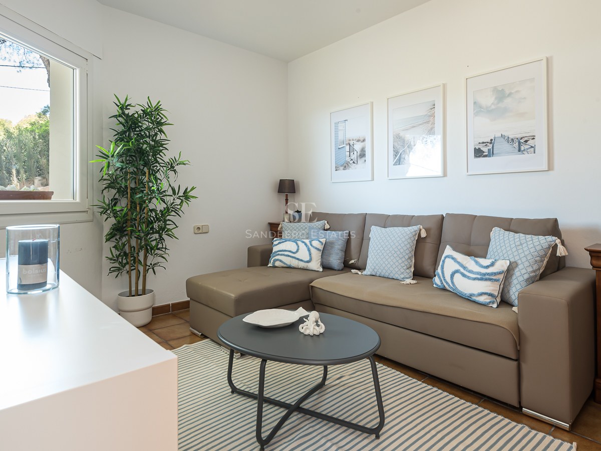Un salon lumineux avec un canapé en L taupe, des coussins bleus, un tapis à rayures et des tableaux sur le thème de la mer.