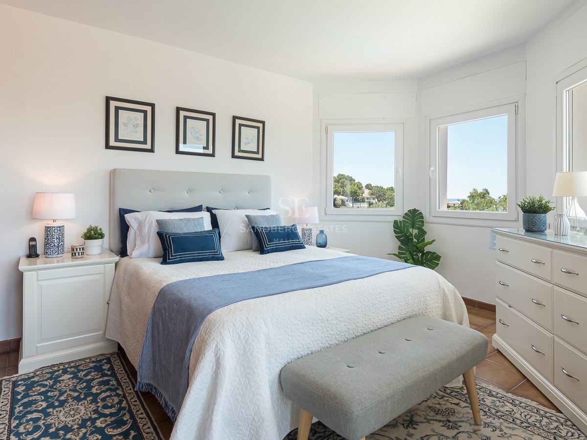 Chambre lumineuse avec lit blanc, coussins bleus, commode blanche, grandes fenêtres et sol en terre cuite.