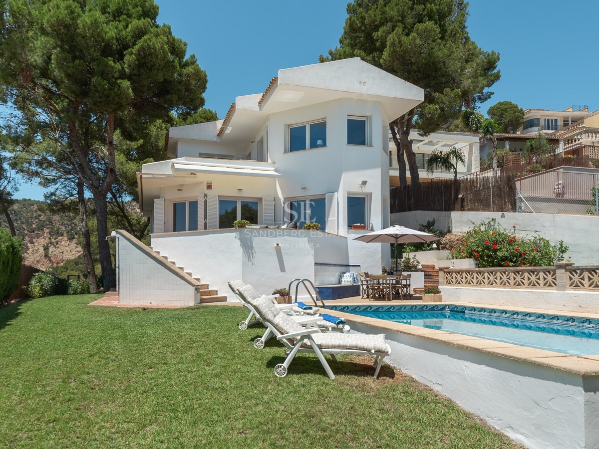 Villa blanche de deux étages avec piscine, transats et pelouse entourée de pins.