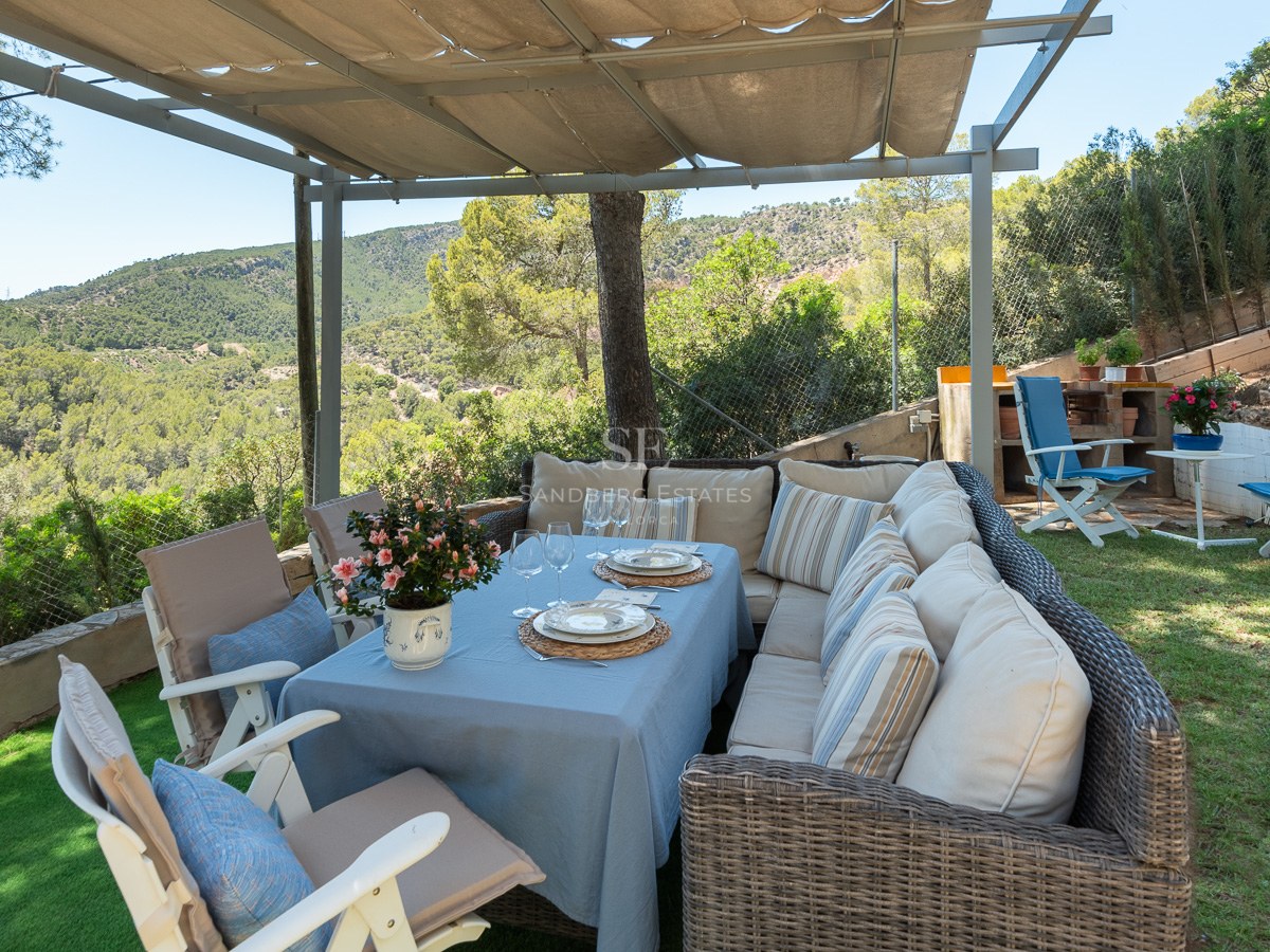 Espace dînatoire extérieur sous pergola avec canapés en osier et vue sur les collines verdoyantes.