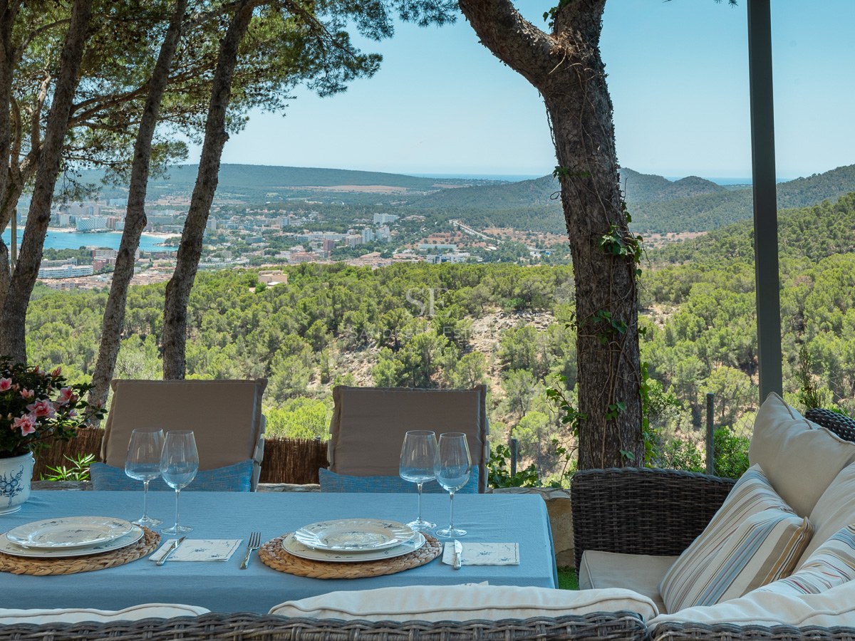 Table à manger extérieure et canapé en osier sur une terrasse ombragée surplombant une ville côtière et la mer.