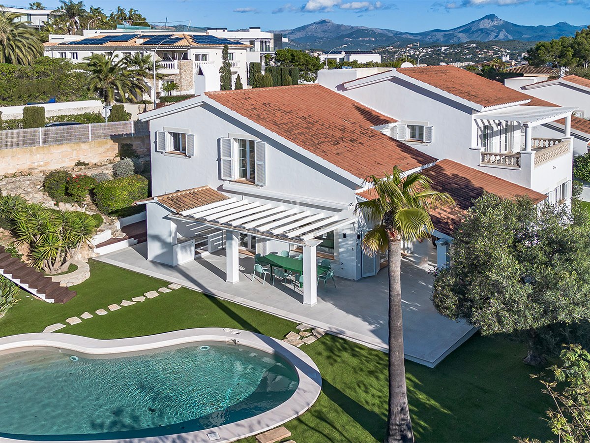 Vue aérienne d'une villa blanche, toit en terre cuite, piscine en forme de rein et jardin sur fond de montagnes.