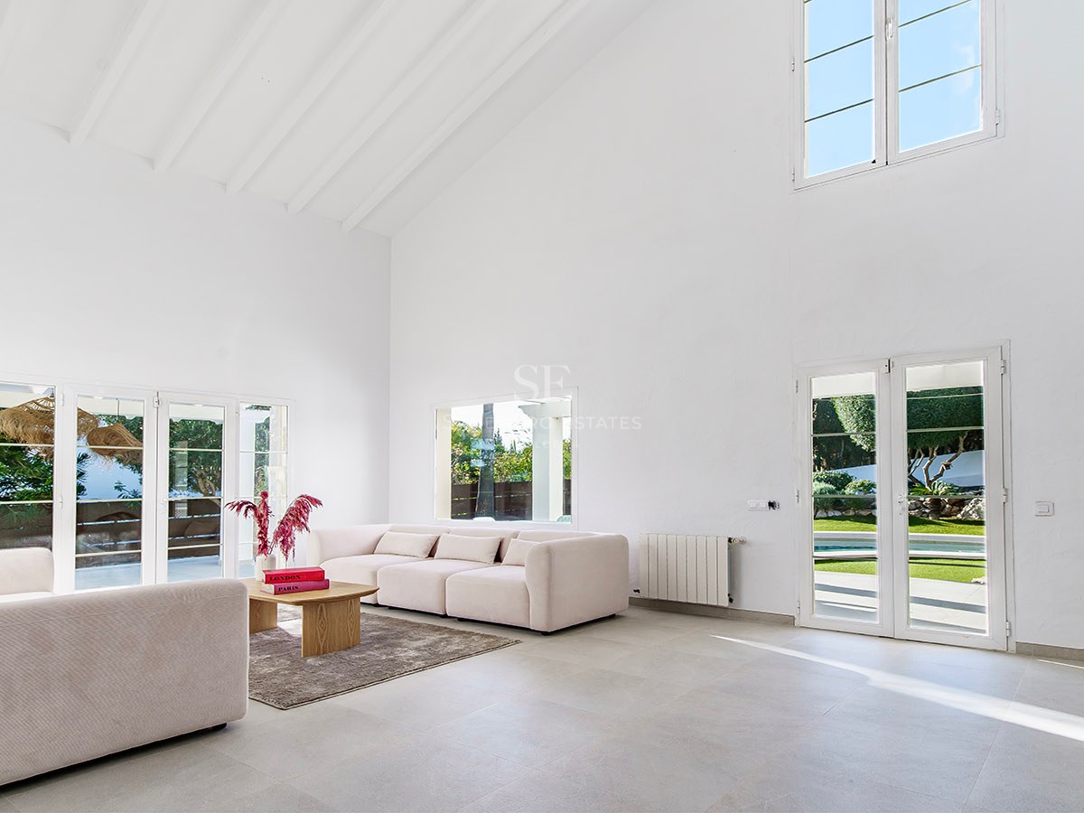 Salon spacieux aux plafonds blancs, canapés beige et portes-fenêtres ouvrant sur le jardin ensoleillé.