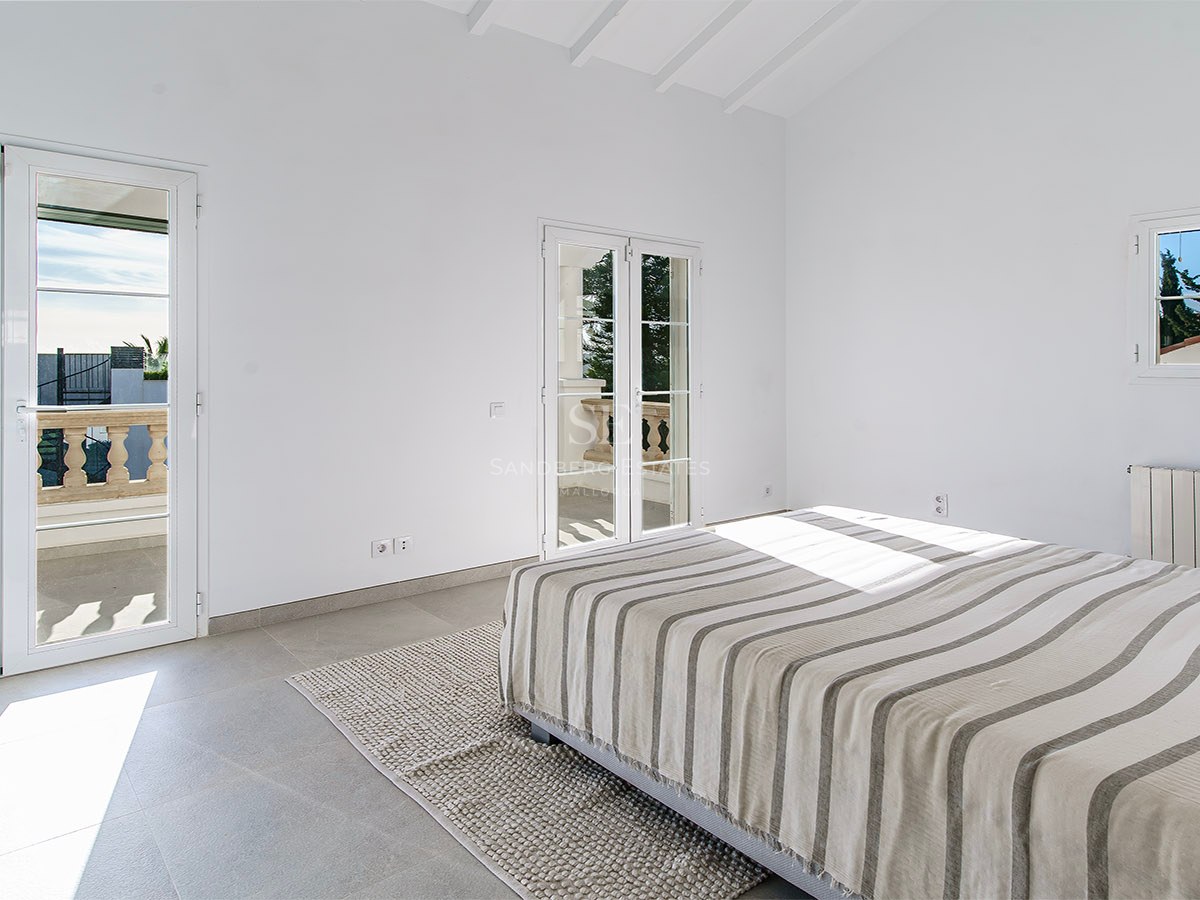 Chambre minimaliste avec lit rayé, sol en grès gris, hauts plafonds à poutres blanches et balcon ensoleillé.