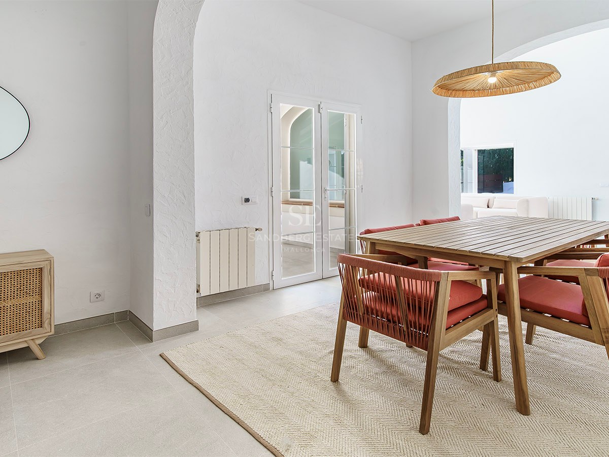 Salle à manger lumineuse avec table en bois, chaises terre cuite, luminaire en osier et arches blanches.