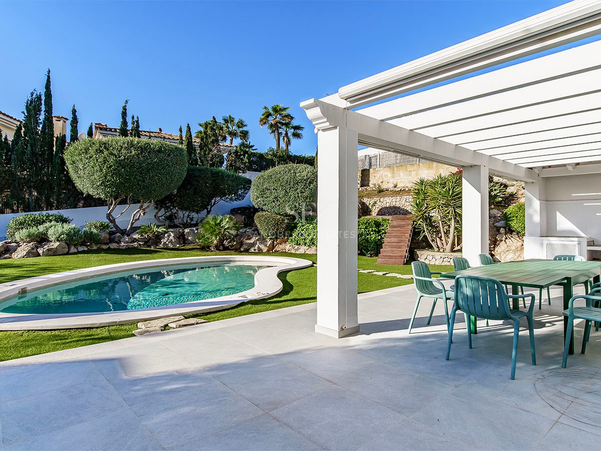 Pergola blanche abritant un coin repas extérieur avec chaises vertes à côté d'une piscine et d'un jardin paysager.