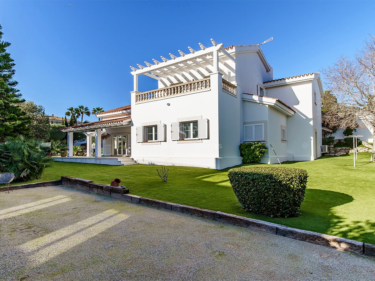Villa blanche à deux étages avec pergola, pelouse soignée et pins parasols sous un ciel bleu intense.
