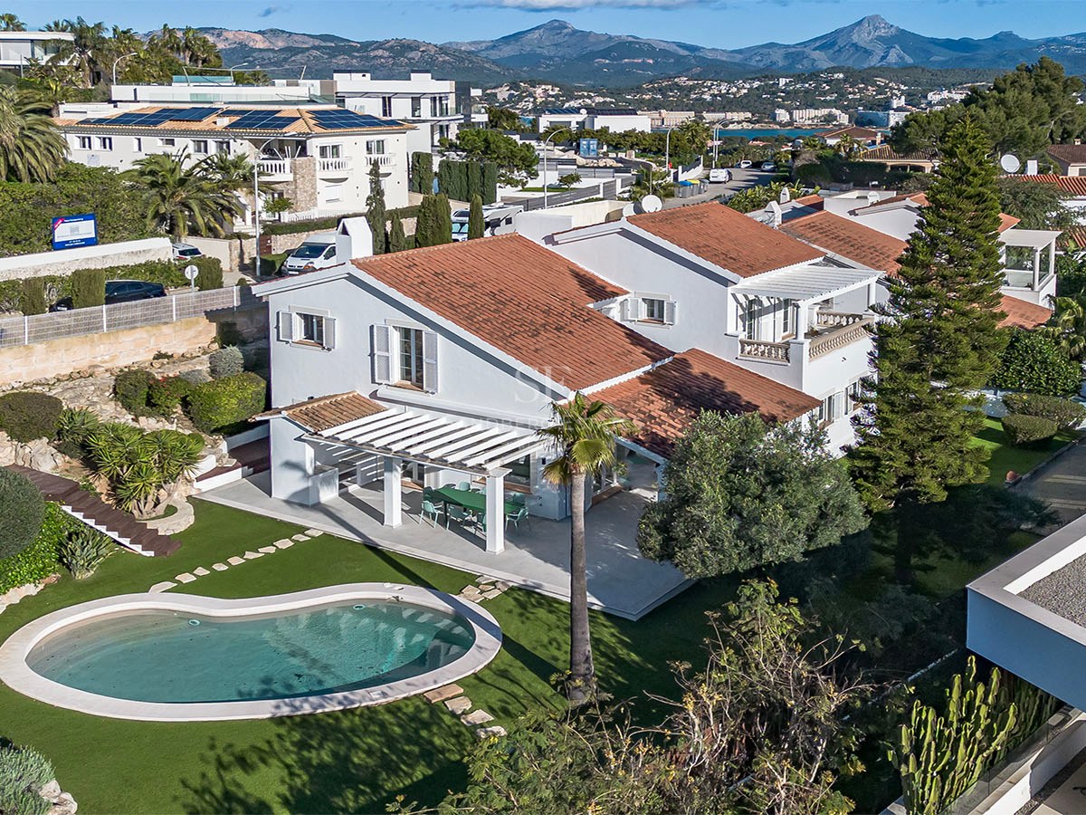 Vue aérienne d'une villa blanche au toit de tuiles avec piscine en forme de haricot et jardin devant les montagnes.