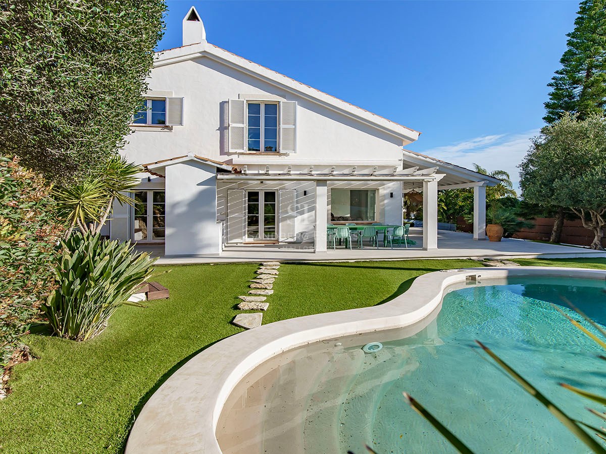 Vue arrière d'une villa blanche avec piscine turquoise, pelouse verdoyante et terrasse ombragée sous un ciel bleu.