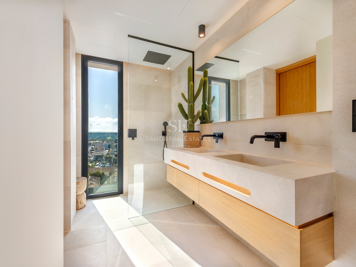 Baño contemporáneo con lavabo doble de piedra, ducha de cristal y ventanal con vistas a la costa.