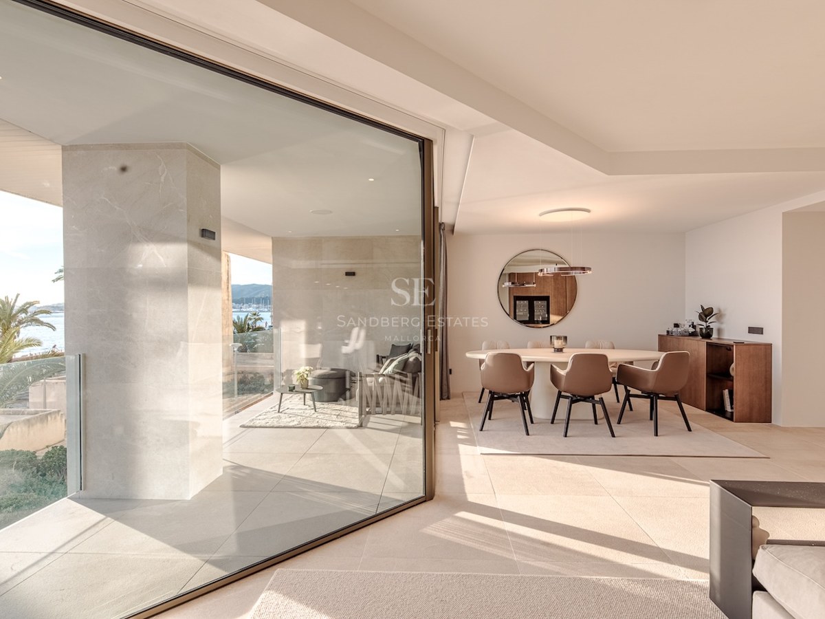 Interior dining room with round table and brown chairs, opening through glass doors to a sunny terrace with sea views.