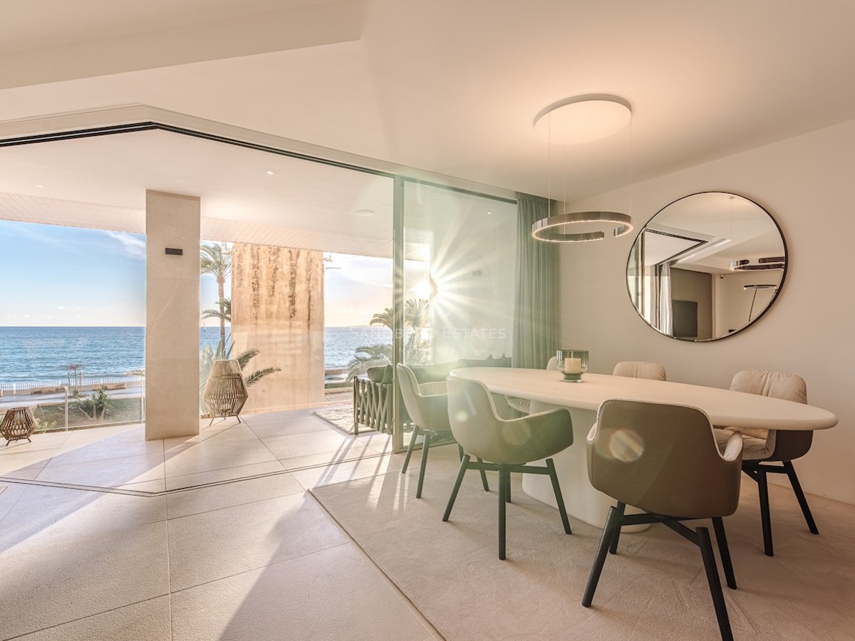 Modern indoor dining area with an oval table and floor-to-ceiling windows opening to a sunlit terrace by the sea.