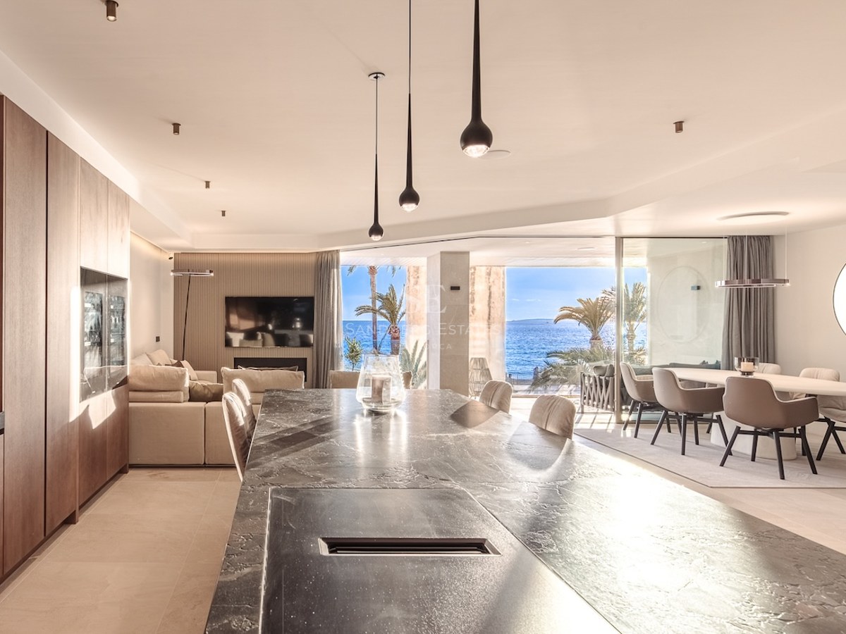 Modern open-plan kitchen and living room featuring a dark stone island, wood cabinetry, and floor-to-ceiling windows overlooking the sea.