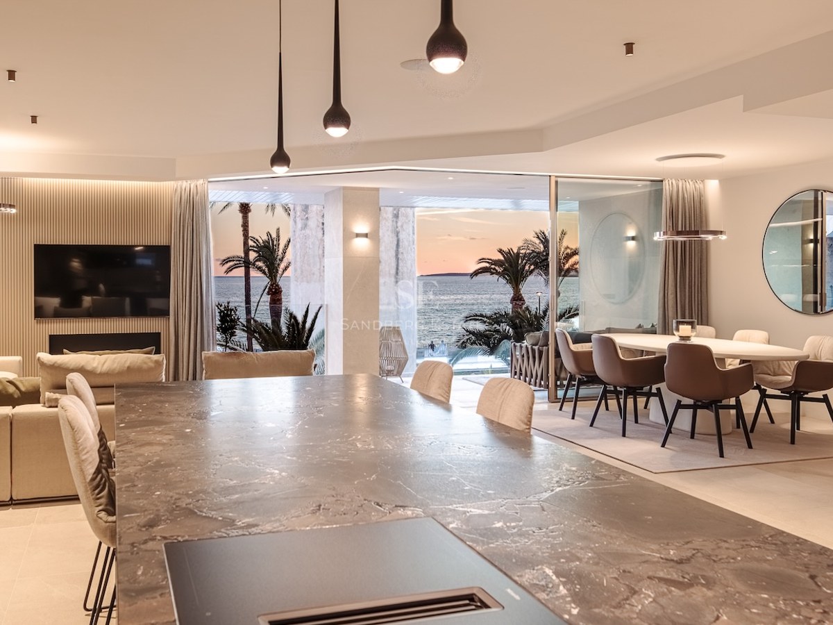 Modern open-plan living and dining space with a marble island, overlooking a terrace with palms and the sea at sunset.
