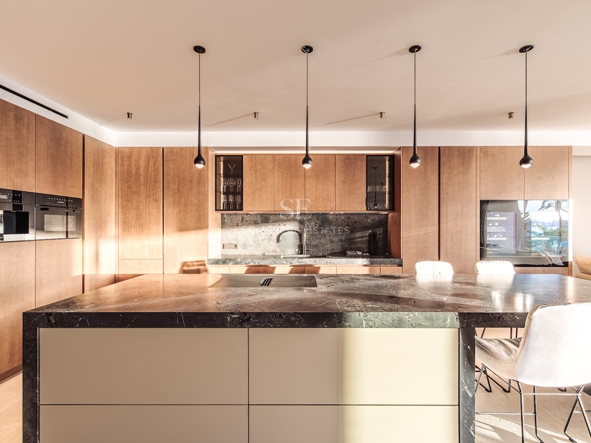 Modern kitchen featuring a large dark marble island with seating, natural wood cabinets, and minimalist pendant lights.