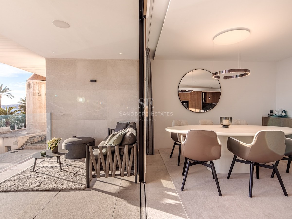 A modern dining room with a white round table and beige chairs, transitioning into an outdoor terrace with sea views.