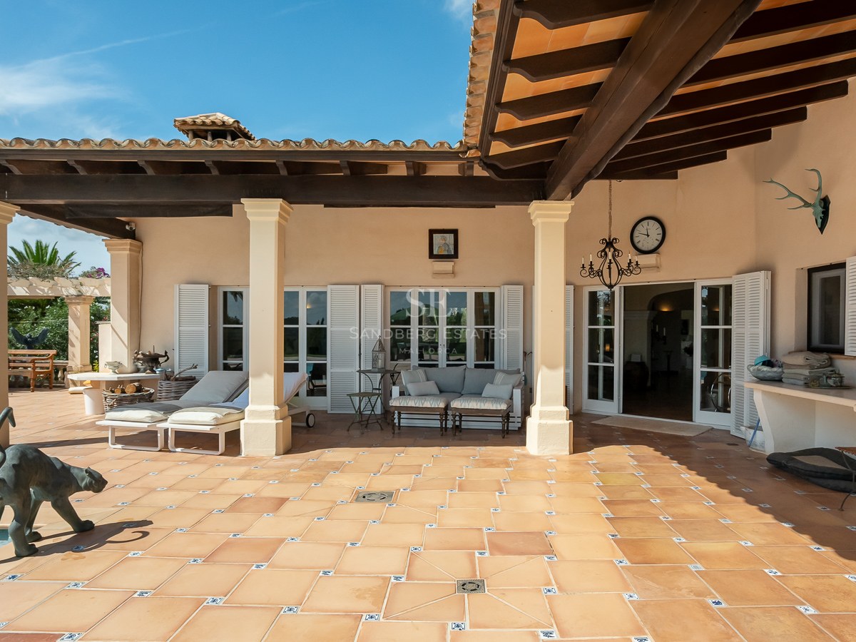 Terrakotta-Terrasse mit dunklen Holzbalken, weißen Säulen, Sonnenliegen und weißen Fensterläden im Sonnenlicht.