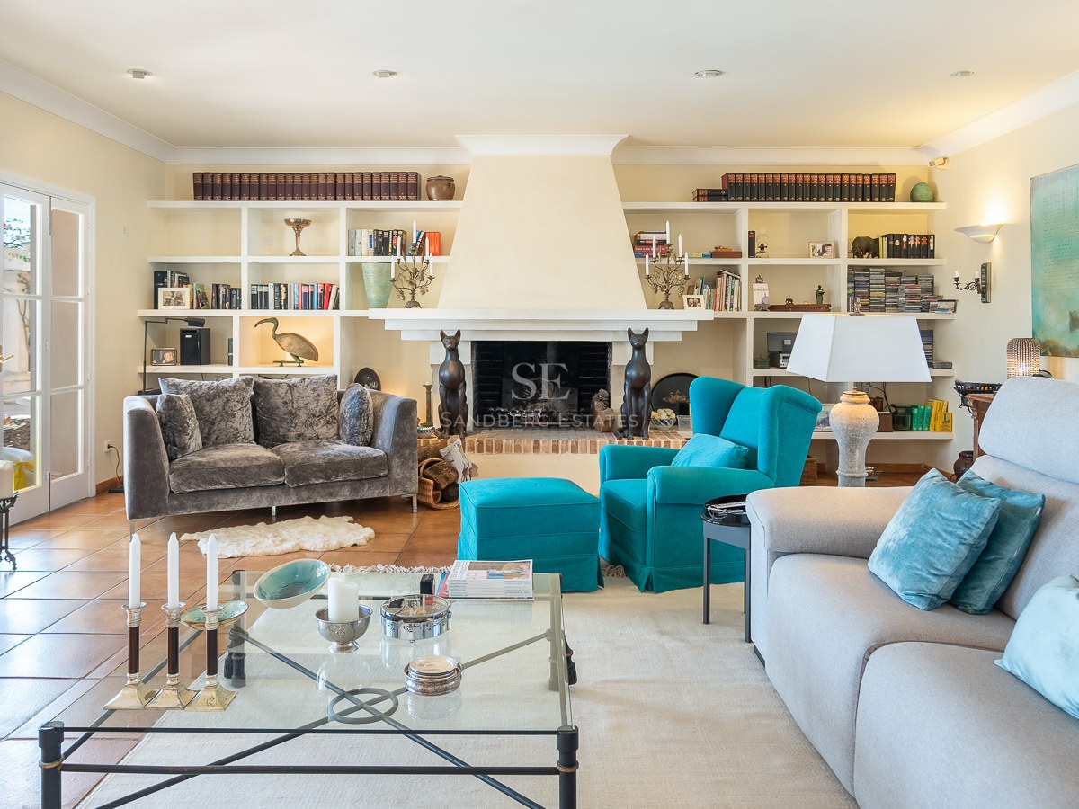Helles Wohnzimmer mit Terrakottaboden, grauen Sofas, türkisfarbenem Sessel, Kamin und Wandregalen.