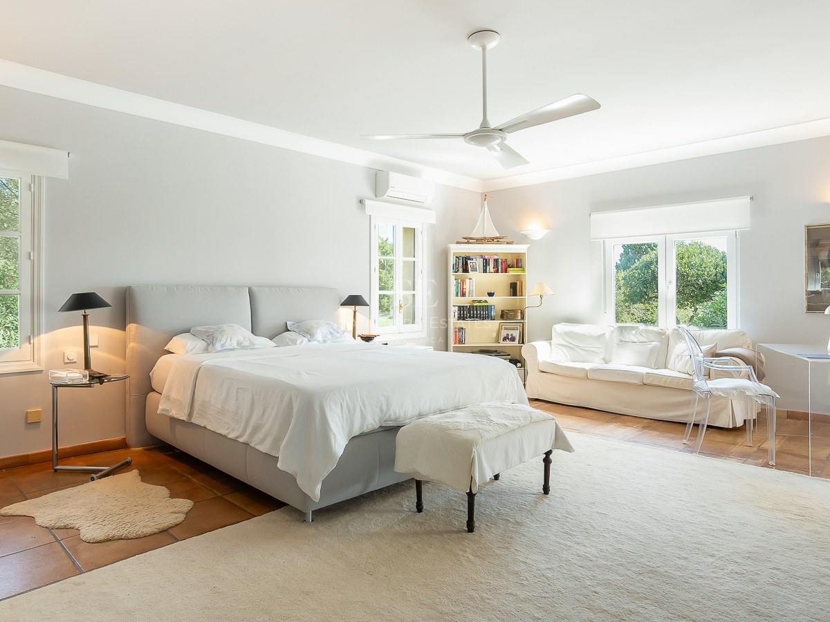 Großzügiges Hauptschlafzimmer mit Terrakottaboden, weißem Sofa, Glastisch und Fenstern mit Blick ins Grüne.