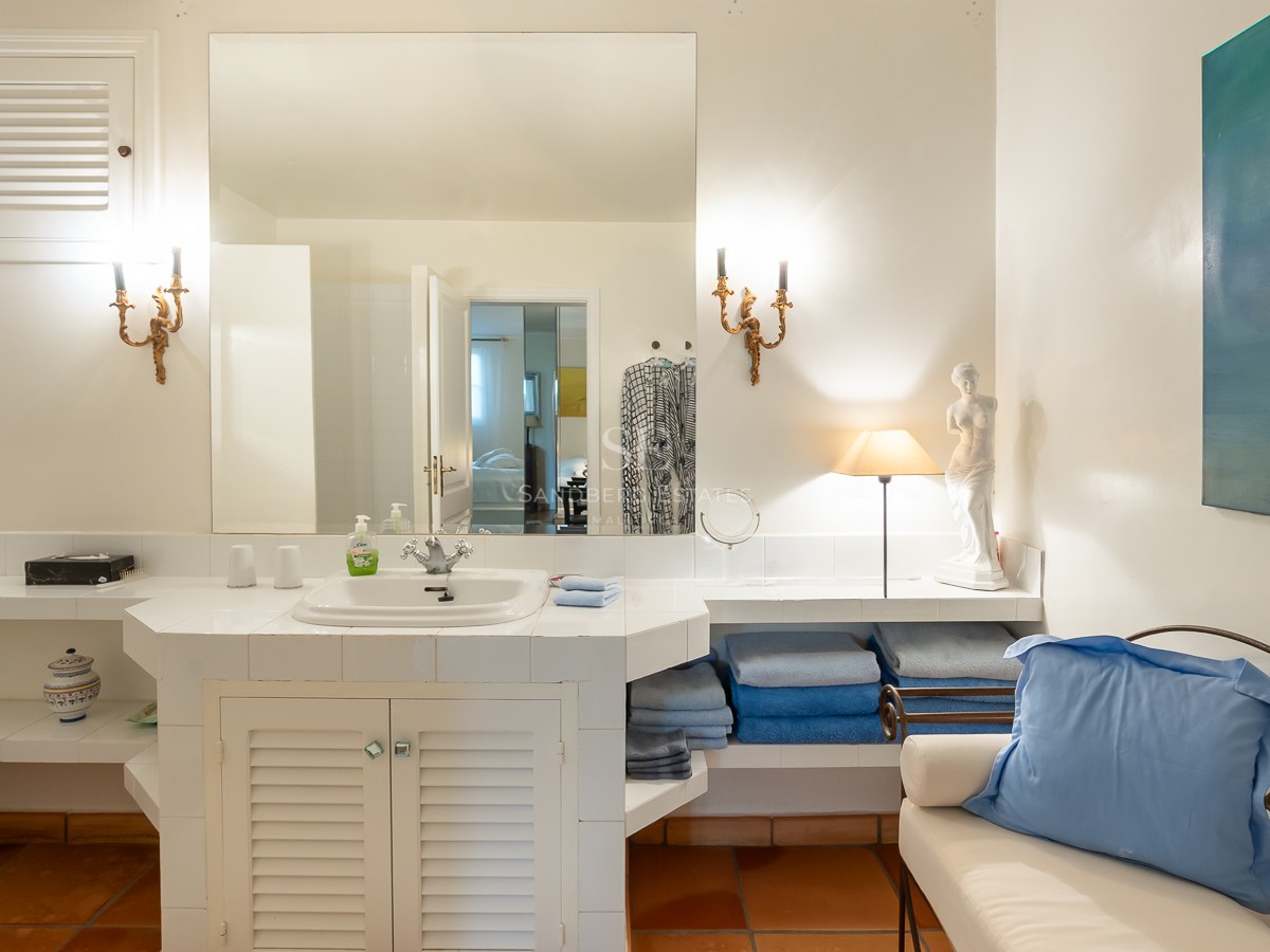 Salle de bain carrelée en blanc avec sol en terre cuite, grand miroir, appliques dorées et touches de bleu.