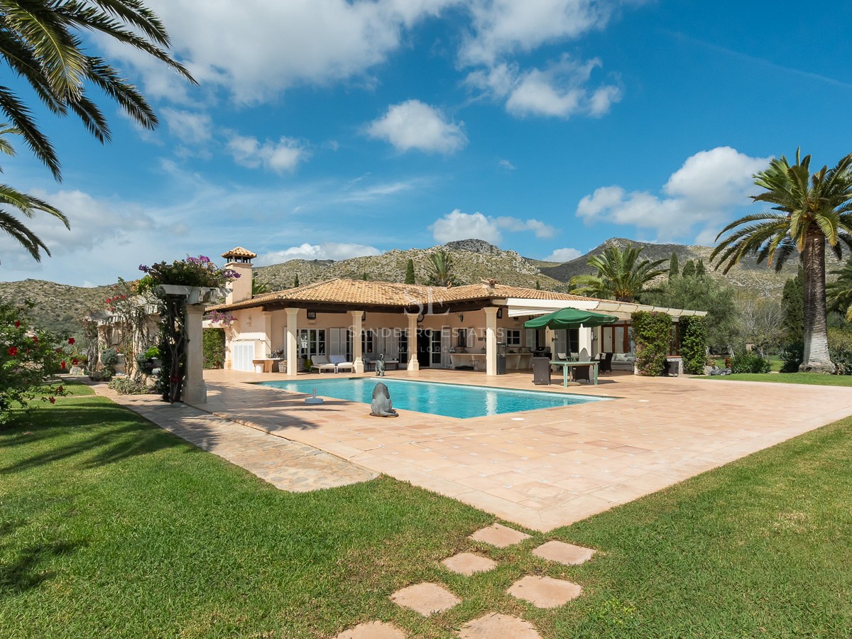 Spanische Villa mit rechteckigem Pool, Terrakotta-Terrasse, Palmen und Bergen im Hintergrund unter blauem Himmel.