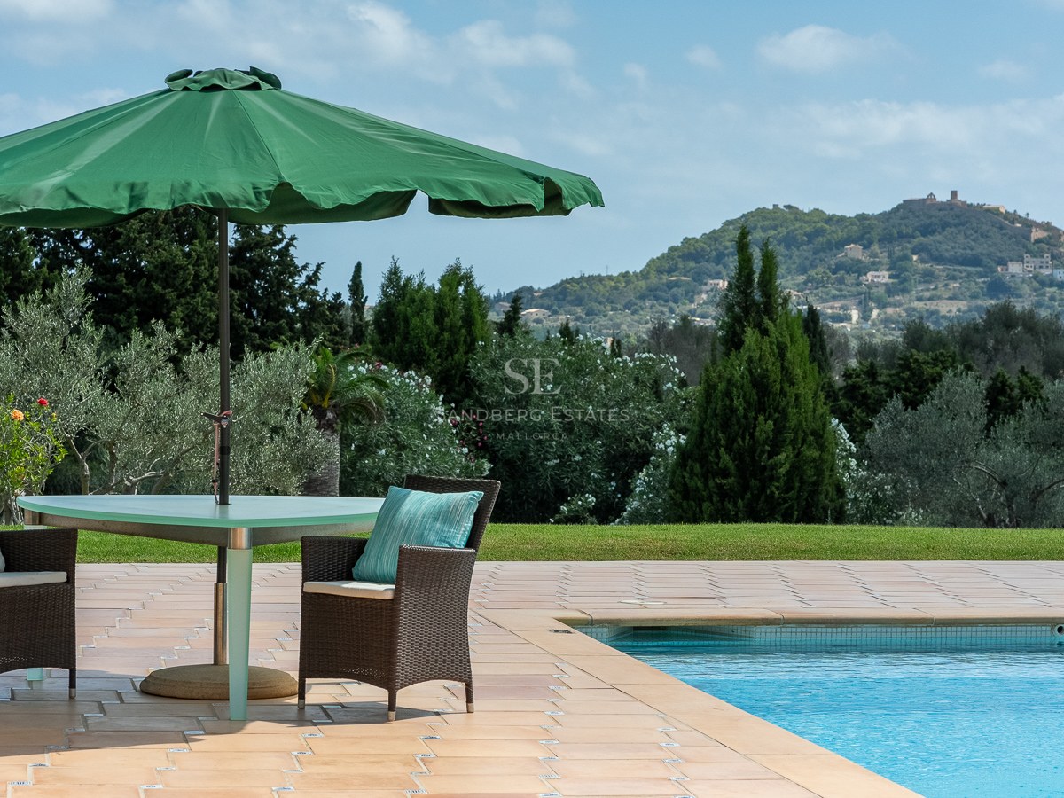 Terrasse au bord de la piscine avec chaises en osier sous un parasol vert, surplombant des jardins et des montagnes.