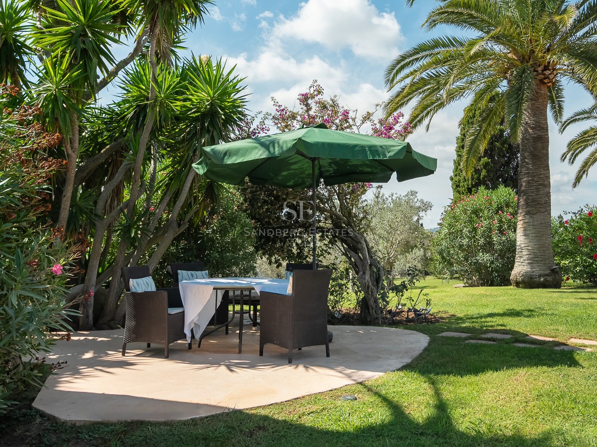 Table à manger ombragée avec chaises en osier sur un patio en pierre, entourée de palmiers et de pelouse verte.