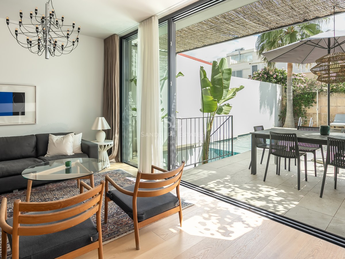 Salon lumineux avec canapé sombre et chaises en bois ouvrant sur une terrasse ensoleillée avec piscine et coin repas.