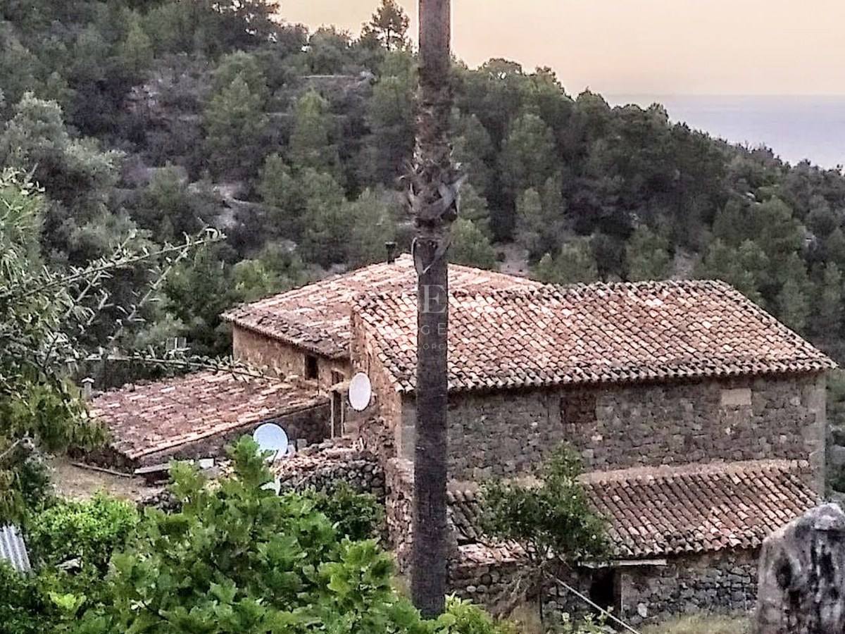 Traditional stone building with terracotta tiled roofs nestled in a lush green hillside overlooking the sea.