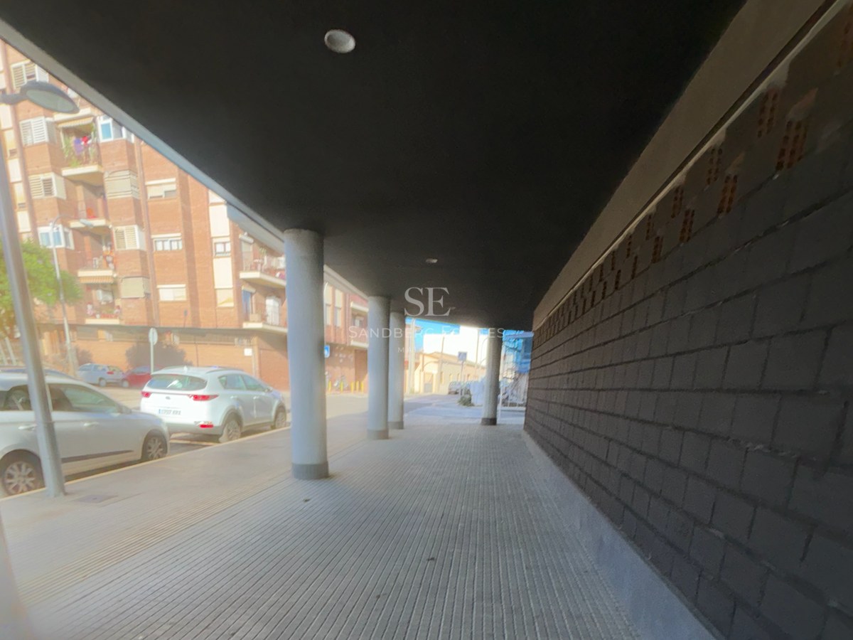 A covered sidewalk with concrete pillars and a dark brick wall next to a city street with parked cars.