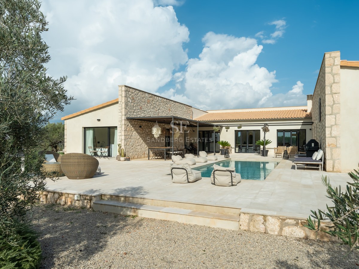 Extérieur d'une villa de luxe avec piscine centrale, murs en pierre naturelle et vaste terrasse pavée sous un ciel bleu.
