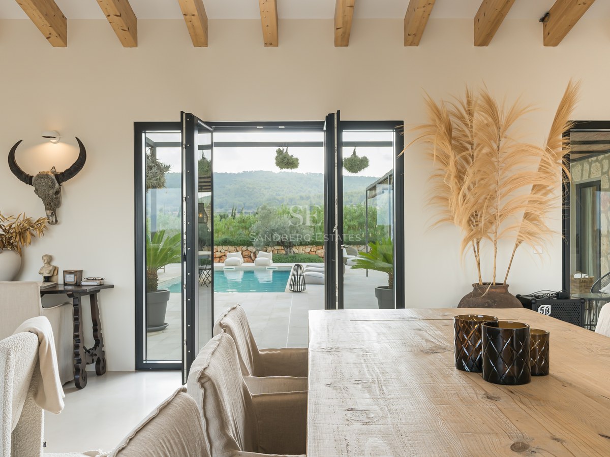 Salle à manger avec table en bois, poutres apparentes et larges baies vitrées donnant sur une terrasse et piscine.