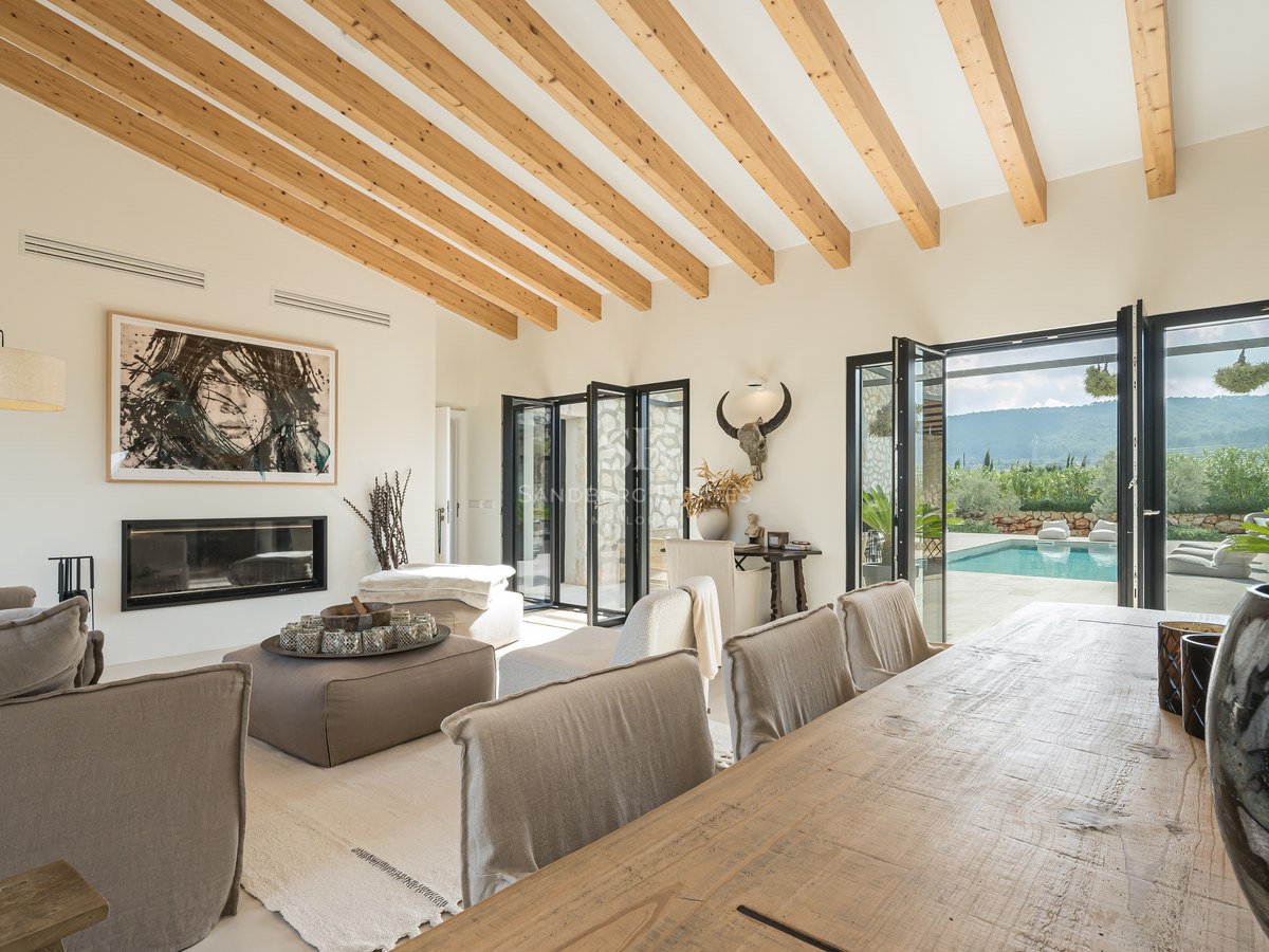 Salon élégant avec plafond voûté en bois, canapés en lin, cheminée et portes pliantes donnant sur la piscine.