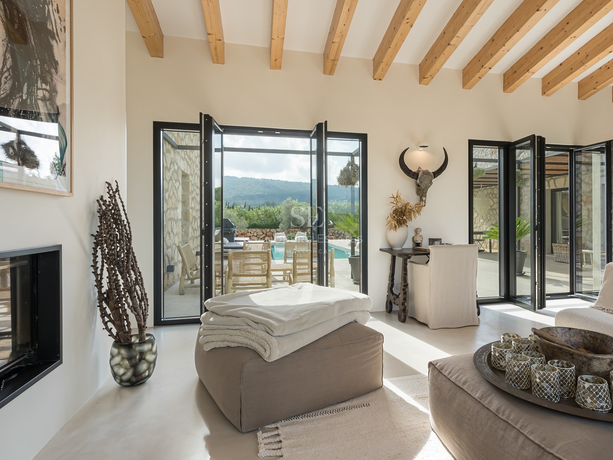 Salon moderne avec hauts plafonds à poutres, portes vitrées pliantes ouvrant sur une terrasse et une cheminée épurée.