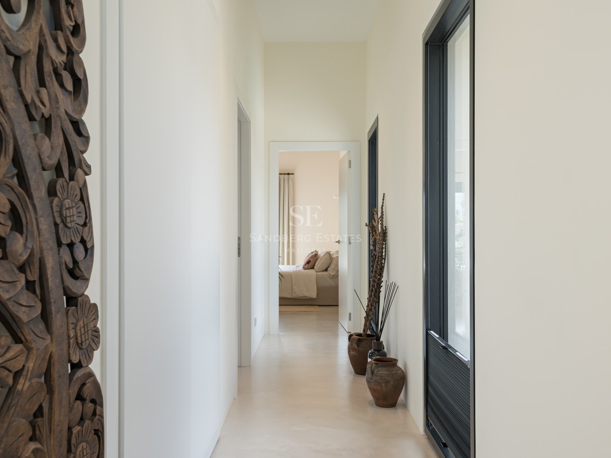 Couloir minimaliste avec sol en micro-ciment, panneau en bois sculpté et vases en terre cuite menant à une chambre.