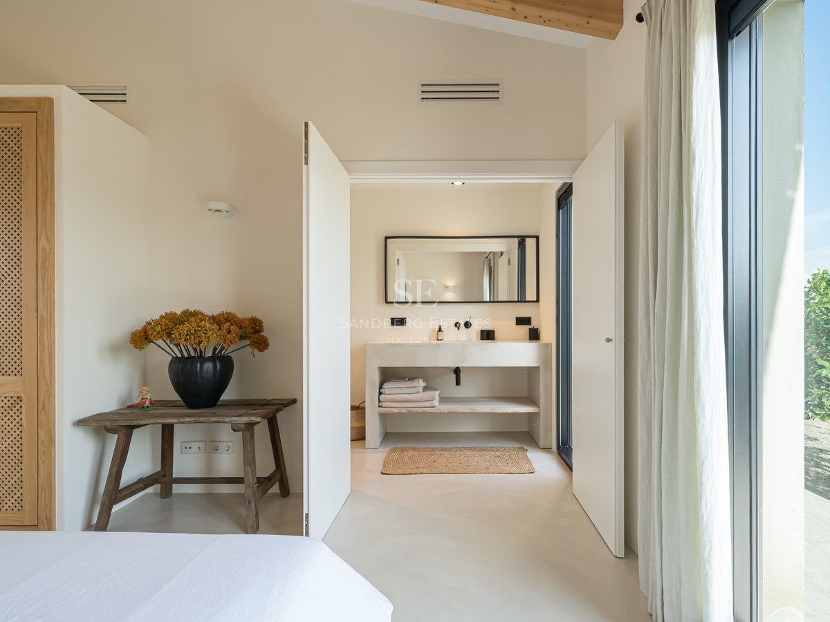 Chambre lumineuse avec poutres en bois et murs blancs s'ouvrant sur une salle de bains moderne en pierre.