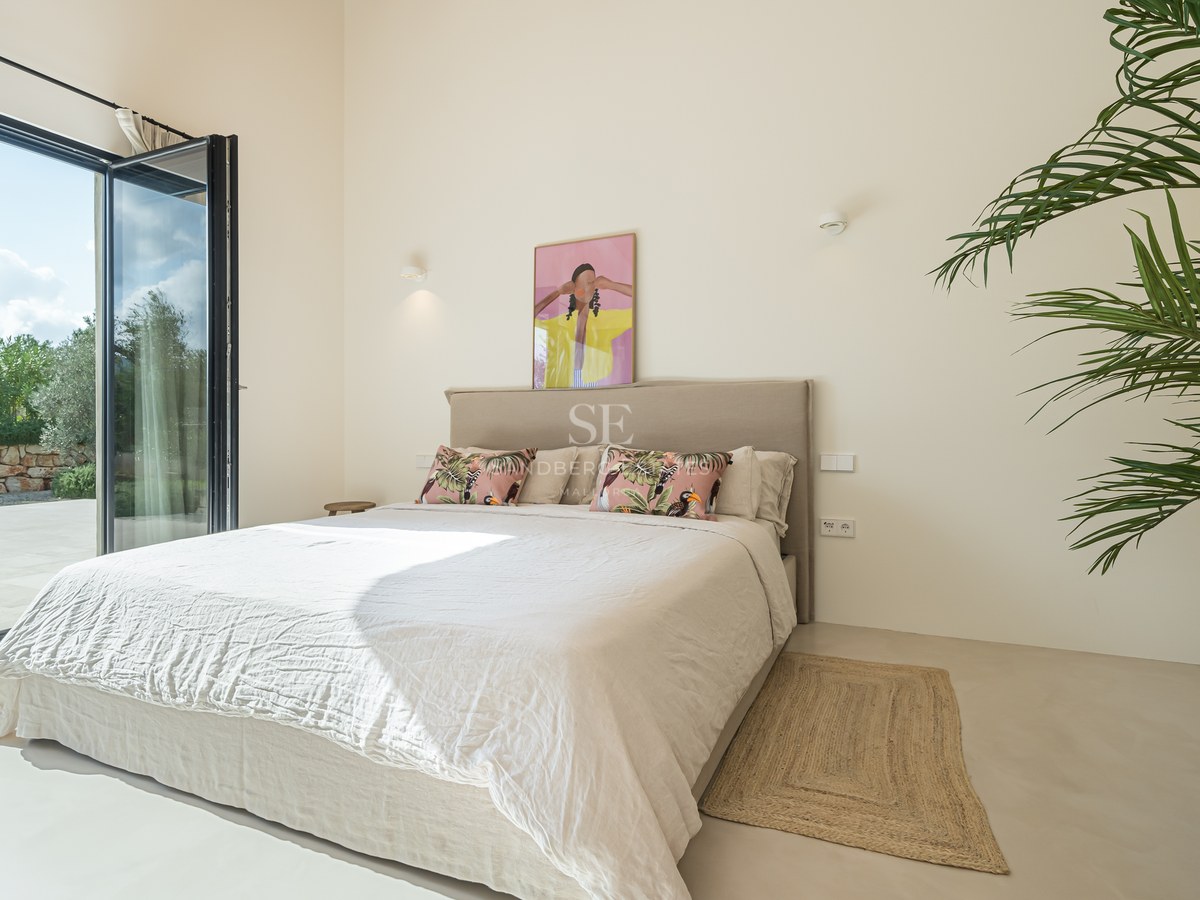 Chambre lumineuse avec baies vitrées ouvrant sur une terrasse, literie neutre et grande plante verte.