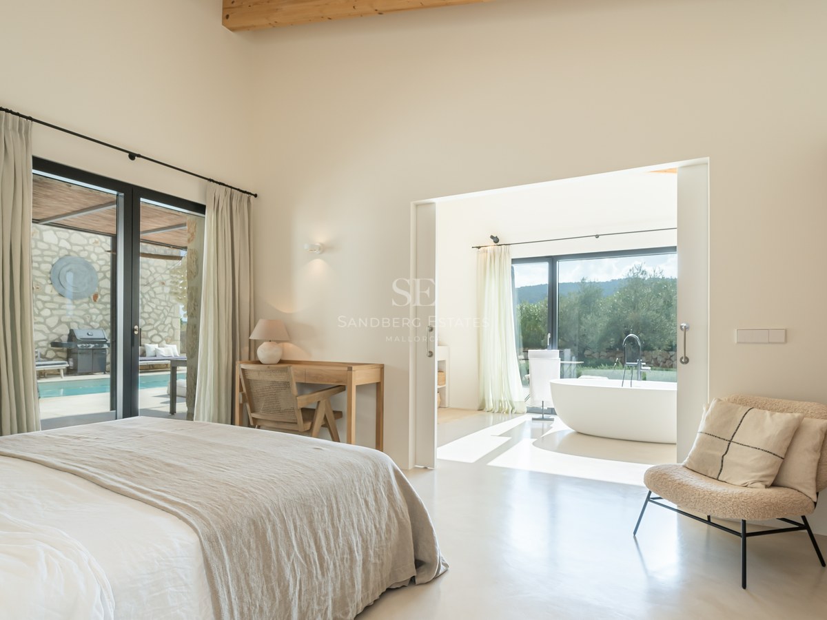 Chambre principale moderne avec poutres en bois, salle de bain attenante, baignoire îlot et accès terrasse.